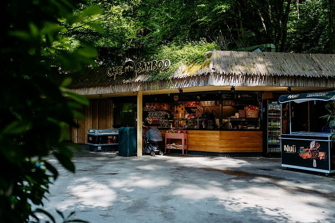 Ein Kiosk mit Stohdach. Rechts und links ist eine Eistruhe zu sehen und mittig eine Frau mit Kinderwagen.