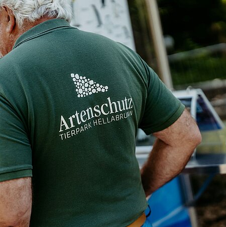 Ein älterer Herr wird von schräg-hinten fotografiert. Er trägt ein dunkelgrünes T-Shirt mit der weißen Aufschrift Artenschutz, welche im Vordergrund des Fotos steht.