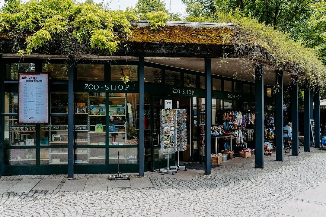 Weitwinkelaufnahme des Zooshops. Links sieht man einen Infomonitor hängen und rechts eine große Auswahl an Plüschtieren und Postkarten.