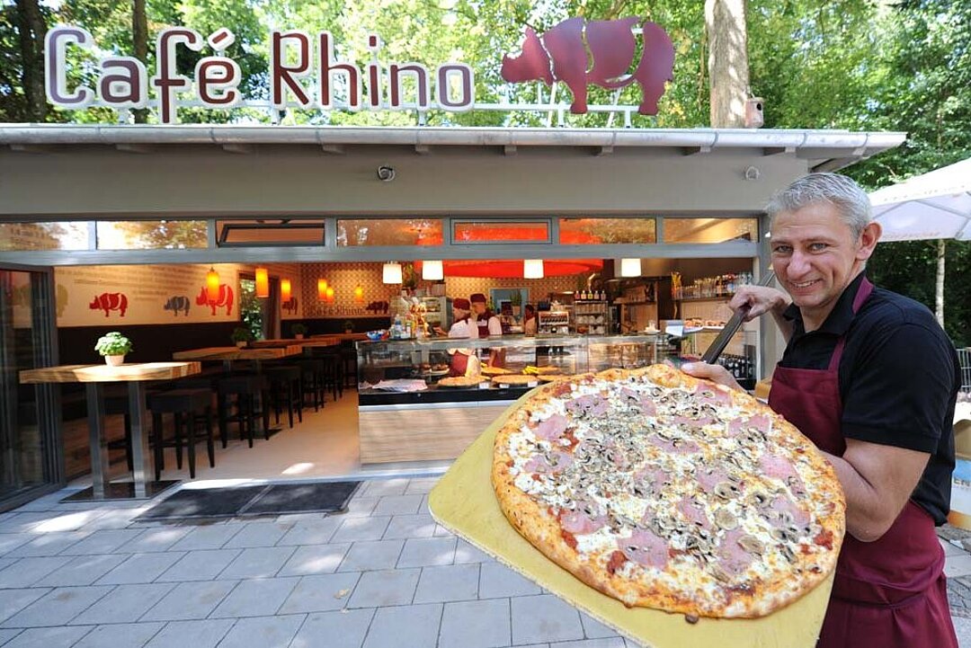 Ein Mitarbeiter des Café Rhinos hält eine große Pizza in die Kamera, hinter ihm die Außenfasade des Cafés.
