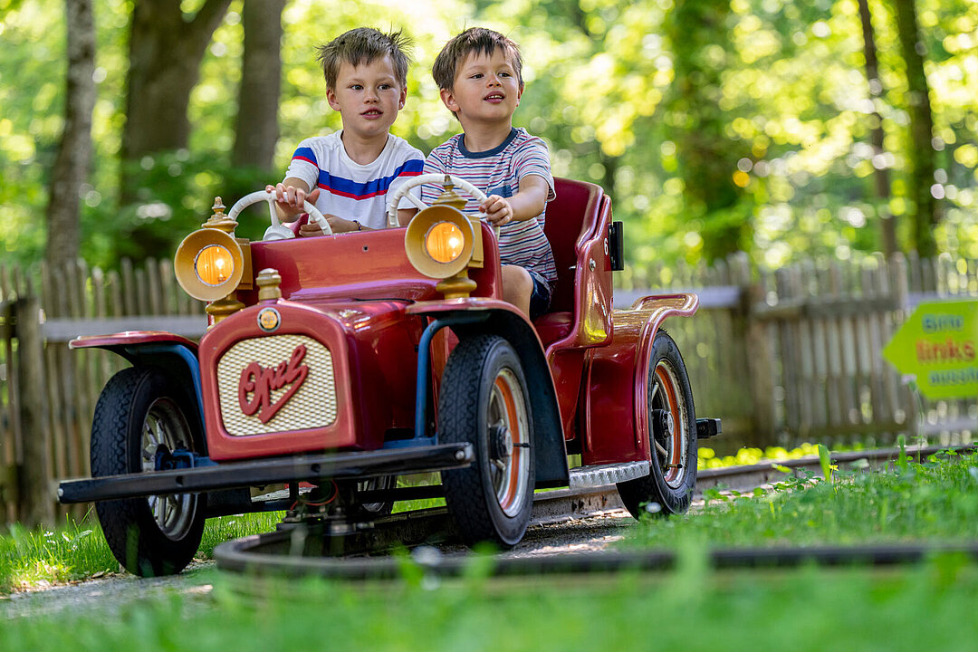 Zwei Kinder im Grundschulalter nutzen die Oldtimer-Bahn und sitzen in einem Auto.